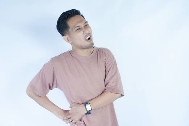 istock A man in a pink shirt bends over slightly and holds his lower back with a pained expression, indicating back pain or discomfort. The plain light blue background emphasizes his distress 2163364146