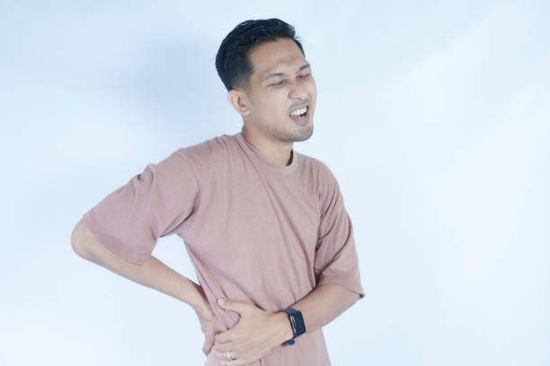 istock A man in a pink shirt bends over slightly and holds his lower back with a pained expression, indicating back pain or discomfort. The plain light blue background emphasizes his distress 2163364170