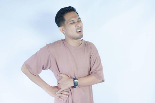 istock A man in a pink shirt bends over slightly and holds his lower back with a pained expression, indicating back pain or discomfort. The plain light blue background emphasizes his distress 2163364183