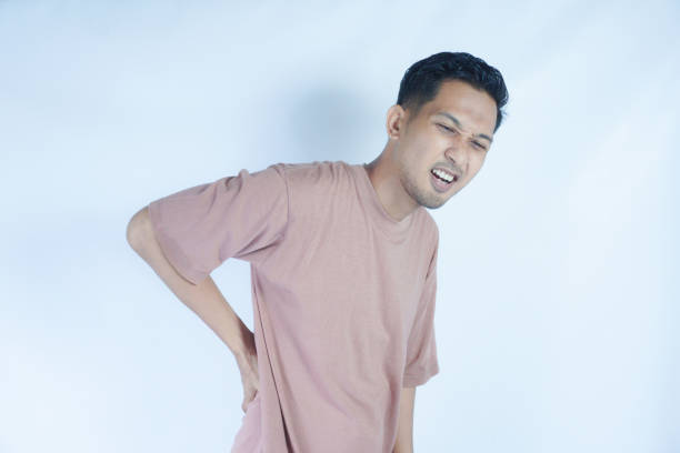 istock A man in a pink shirt bends over slightly and holds his lower back with a pained expression, indicating back pain or discomfort. The plain light blue background emphasizes his distress 2163364189