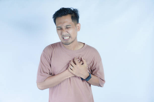 istock A man in a pink shirt clutches his chest with both hands, showing a pained expression, possibly indicating chest pain or discomfort. The plain light blue background emphasizes his distress 2163364363