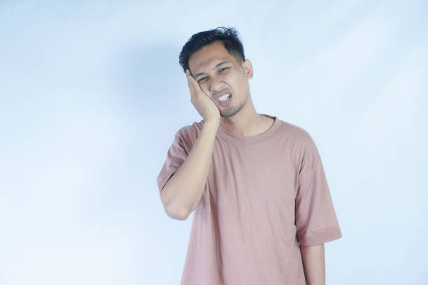 istock A man in a pink shirt holds his jaw with a pained expression, indicating a toothache or discomfort. The plain light blue background emphasizes his distress and highlights his posture and expression. 2163364402