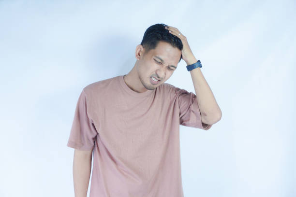 istock A man in a pink shirt holds his head with a pained expression, indicating a headache or discomfort. The plain light blue background emphasizes his distress and highlights his posture and expression. 2163364422