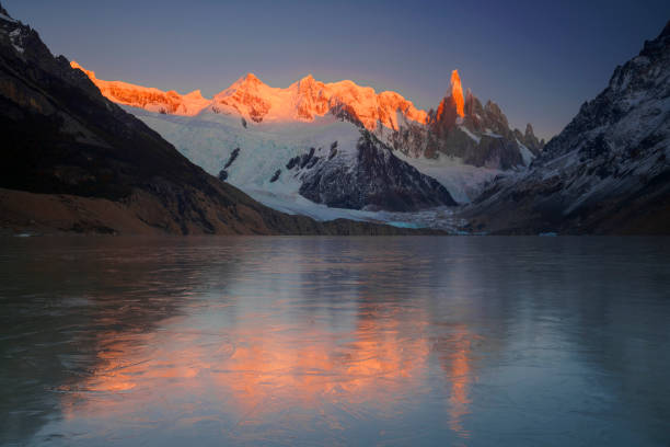 Snow-capped peaks bask in the warm glow of sunrise, creating a striking orange hue against the blue sky. A calm lake at the base of the mountains reflects the majestic scene, enhancing the serene beauty of this natural landscape of El Chaltén, in Patagonia.