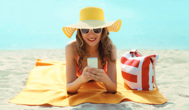 Summer vacation, tourism, happy woman with phone, tourist hat, holding smartphone lying on the beach stock photo