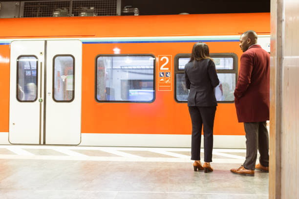 Two professionals stand on a subway platform in the evening, waiting for an orange commuter train to arrive