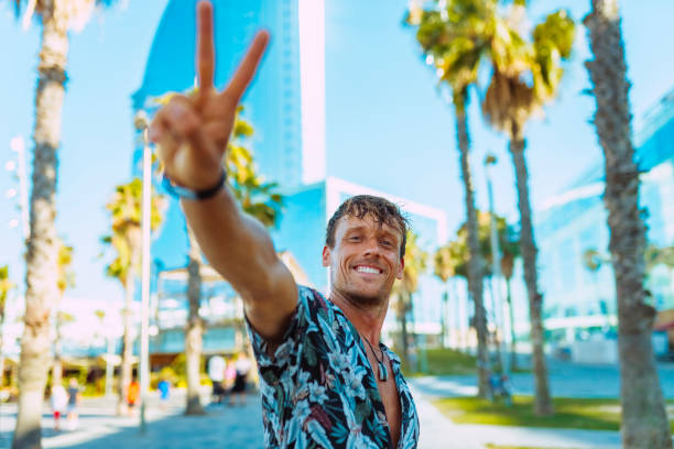 Young stylish man in floral hawaiian shirt, broadly smiles, does peace victory sign at camera at city  outdoors, surrounded by palms at summertime.