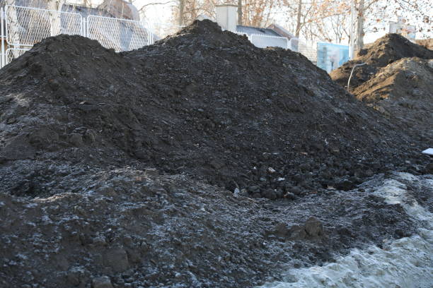 A mound of black soil from road construction sits prominently on the site. The soil, rich and dark, has been excavated and piled up as part of the roadwork process. This pile of soil is a crucial element in preparing the ground for the next phases of construction, such as grading and laying down the roadbed. The surrounding area might show other signs of construction activity, like machinery or partially completed sections of the road. The background features the horizon, which may include distant views of the landscape or ongoing construction efforts. The contrast between the dark soil and the open expanse of the horizon highlights the scale and scope of the road construction project, emphasizing the transition from raw land to developed infrastructure.