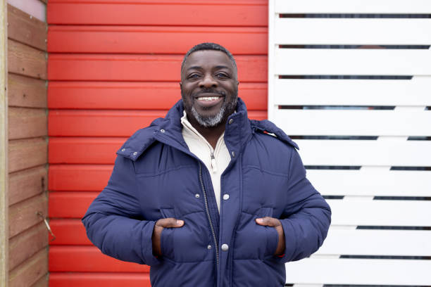 Medium portrait shot of a man standing in front of red and white wood. He is looking directly at the camera and smiling while wearing casual clothing. He is a polio survivor and he lives with disabilities.Videos are available similar to this scenario.