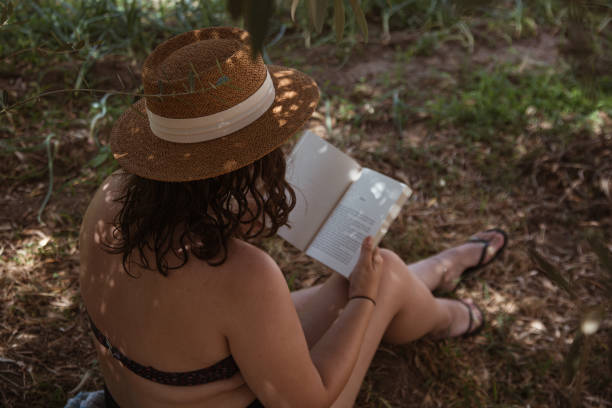 Rear view of a young woman reading a book sitting in the shade of a tree.