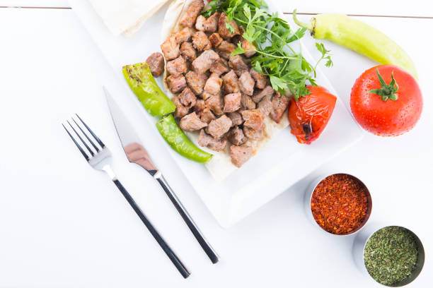 Grilled meat, vegetables and spices on a white plate placed on a white table. The plate contains fried meat cubes, green peppers, parsley and a piece of pita bread. On the right of the plate are a red pepper, a tomato, red pepper flakes and dried herbs. On the left are a fork and a knife. The background is white.