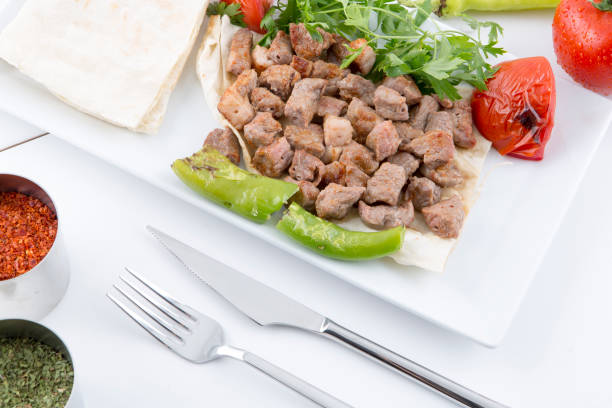 A white plate shows small pieces of meat placed on thinly sliced ​​buttered and toasted bread slices. The pieces of meat look delicately fried and delicious. They are decorated with bright green pepper slices and bright red tomato paste around them. A fork and knife are placed next to the plate. Also included in the photo are a small bowl of red pepper and another bowl of dried herbs. All these details create an eye-catching food presentation.