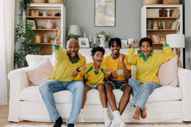 A very happy family of four wearing yellow team jerseys sits on a white couch, enthusiastically raising their arms and cheering together in their bright and cozy living room.