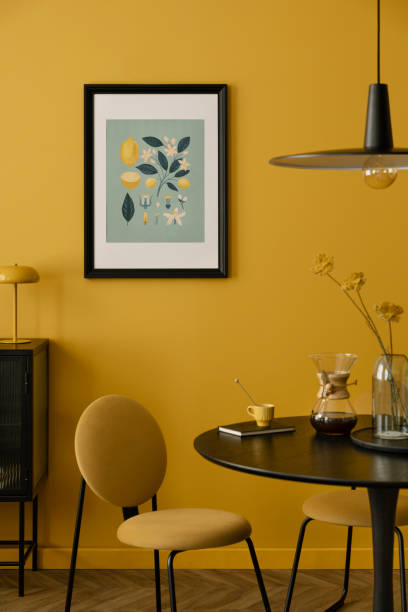 istock Stylish composition of warm dinning room interior with mock up poster frame, round table, yellow chair, glass vase with dried flowers, lamp, and personal accessories. Home decor. Template. 2163425807