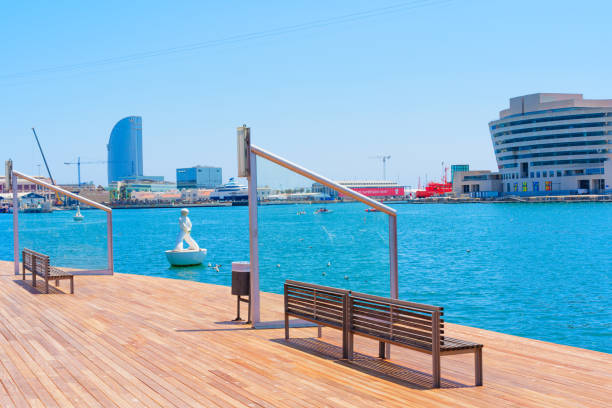 Barcelona, Spain - July 16, 2024: Inviting relaxation zone featuring benches on a wooden pier offering stunning view of modern architecture in Barcelona's port.