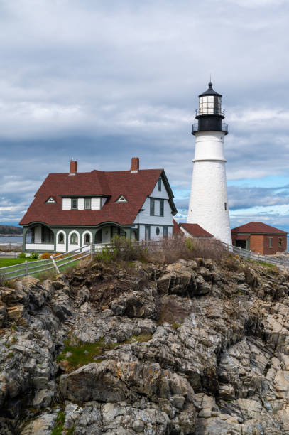 A white lighthouse stands on a rocky cliff overlooking the ocean, with a gray and white house in the foreground.