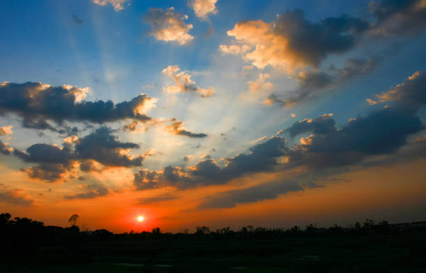 Dramatic sunset sky with vibrant orange and blue hues over lush green rural countryside landscape and silhouetted trees. Peaceful and serene scenic nature view at dusk.