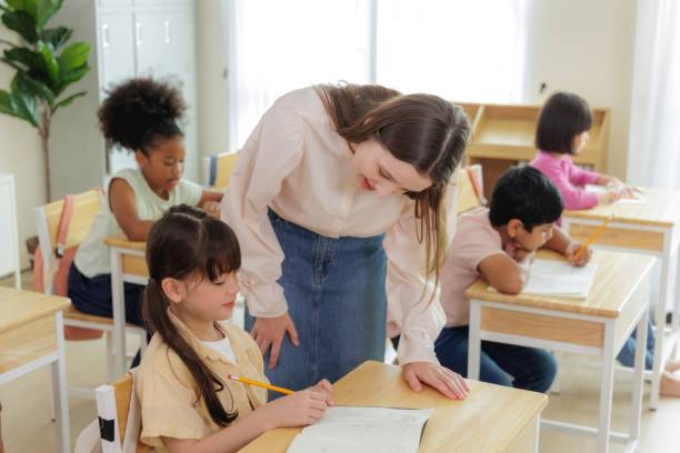 istock A teacher assists a young student in a classroom setting, while other students work at their desks. 2163461124