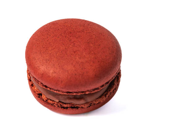 A single, red macaron with a strawberry filling is shown in a close-up view. The macaron is isolated on a white background, which highlights its vibrant red color and textured surface.