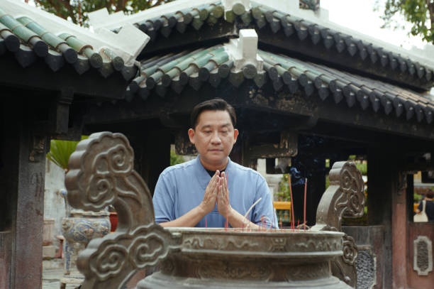 Asian man praying near the cup with aroma sticks while visiting Buddhism temple