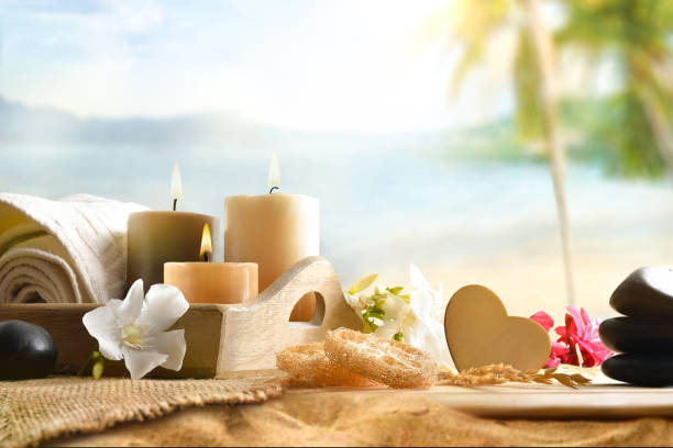 Body and mind wellness with products and decorative elements on a wooden table with candles, towel, and flowers with beach in the background. Front view.