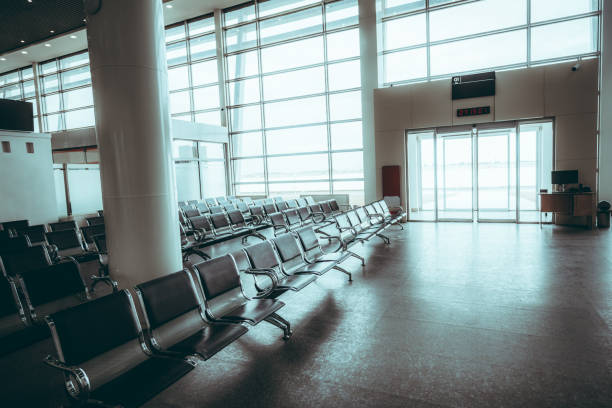 A spacious, empty airport waiting area with rows of metal chairs arranged neatly. Large floor-to-ceiling windows flood the space with natural light, creating a bright, airy atmosphere