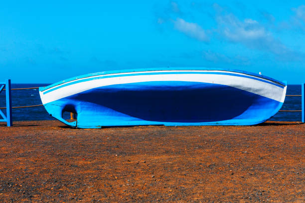 Blue and white boat is sitting on the beach. Boat is not in use and is leaning against a fence