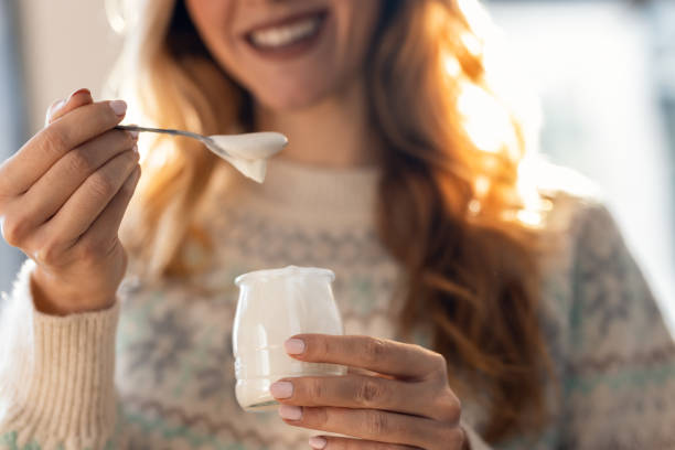 Happy beautiful woman eating yogurt while standing in living room at home Close up of happy beautiful woman eating yogurt while standing in living room at home yogurt stock pictures, royalty-free photos & images