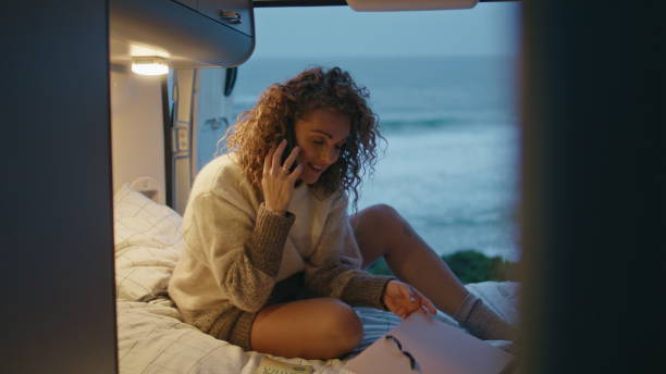 istock Positive freelancer talking cellphone in evening campervan at seashore. 2163510952