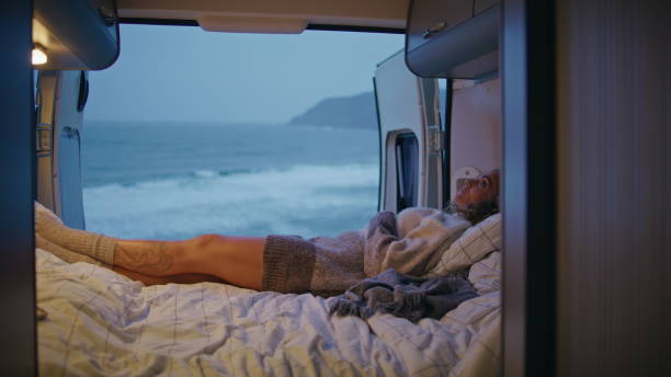 istock Woman enjoy cozy camper van bed spending weekend evening on gloomy seashore. 2163511043