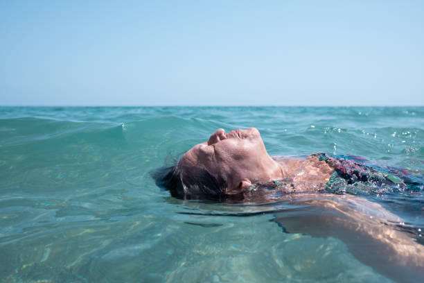 senior white woman with gray hair with her eyes closed and breathing in the sea water in summer. mindfulness and wellbeing.floating in the surface of the water drifting. senior white woman with gray hair with her eyes closed and breathing in the sea water in summer. mindfulness and wellbeing.relaxation and a sense of freedom.mental health and emotional balance. woman cold water swimming stock pictures, royalty-free photos & images