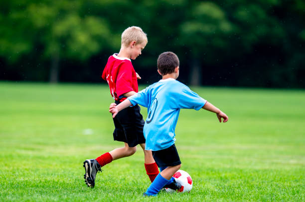 Two children are seen playing soccer against each other as one child winds up for a big kick.  They are wearing red and blue jerseys and focused on the game.
