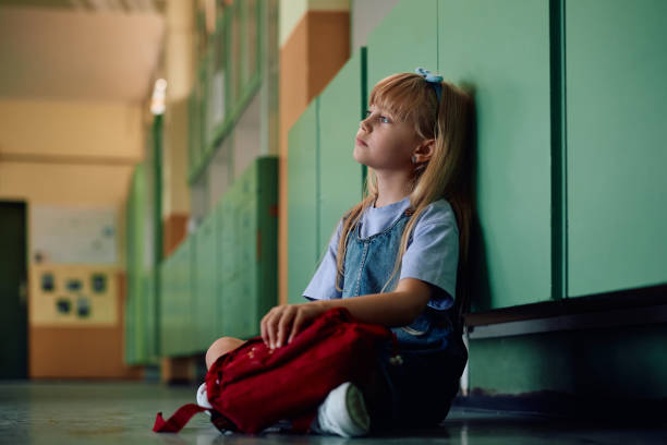 Pensive little girl sitting alone at school hallway. Sad schoolgirl sitting in hallway at elementary school and thinking of something. Copy space. child sadness stock pictures, royalty-free photos & images