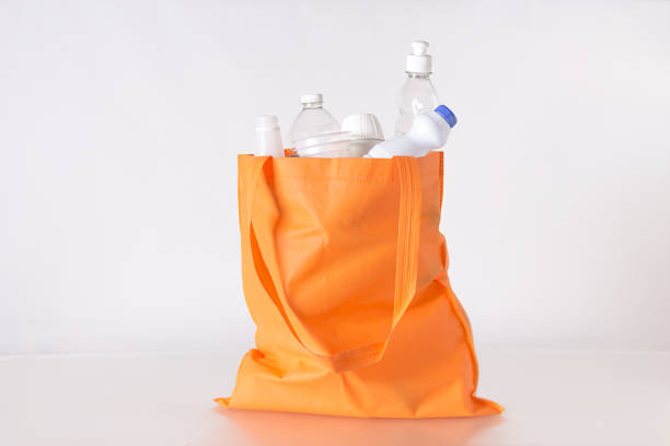 A reusable orange cloth bag filled with recycled plastic bottles of different types on a white surface with high key lighting.