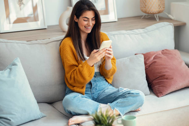 istock Smiling young woman using her mobile phone while sitting on couch at home 2163566988