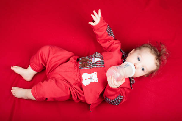 A young child with blond hair, wearing a red and white plaid shirt and a blue hoodie, is captured looking over their shoulder, holding a baby bottle. The warmth of the café setting suggests a casual, comforting atmosphere while natural light floods in from the background, highlighting the toddlers innocent expression.