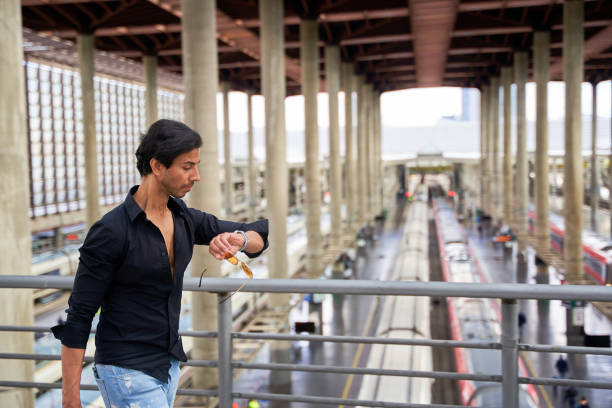 Man in a black shirt and ripped jeans checking his watch at a train station, holding sunglasses, with trains and platforms in the background.