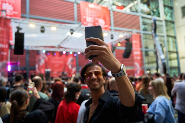 Indian man in black shirt and sunglasses taking a selfie at a concert, smiling amidst a crowd.