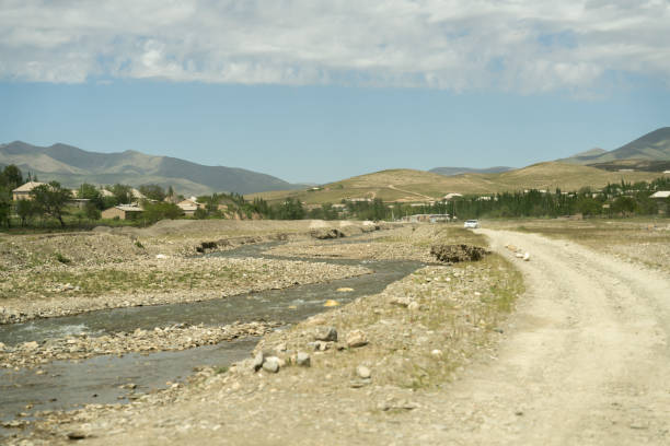A road runs through a desert with a river running alongside it. The road is rocky and dusty, and there are no buildings in the distance. The sky is clear and blue