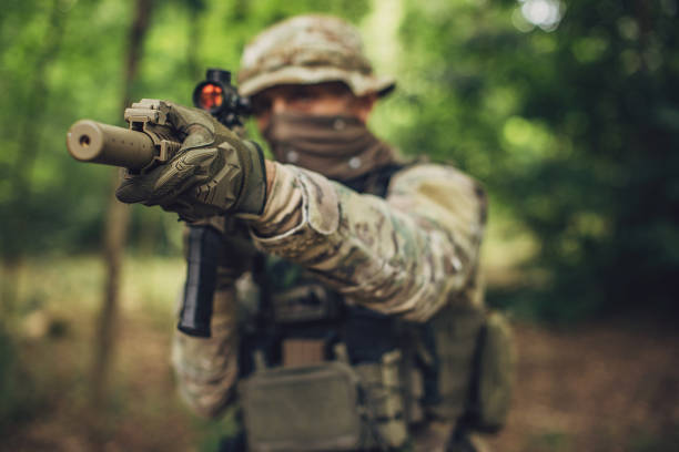 Military soldier dressed in full camouflage aiming a rifle with a tactical visor in a dense forest setting, exemplifying stealth and precision in a natural combat zone.