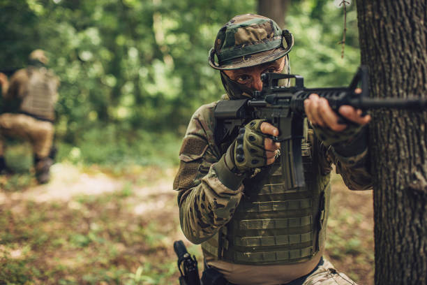 istock Armed soldier in camouflage aiming rifle behind a tree 2163601507