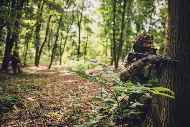 A soldier in camouflage hide behind a tree in a forest, aiming his rifle forward, suggesting a strategic position, with the background portraying a dense, natural setting.