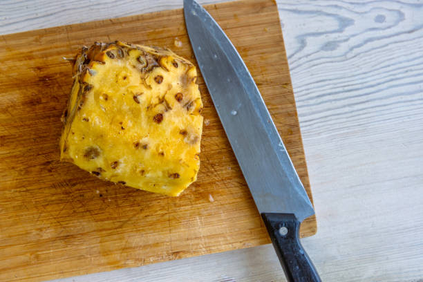 A partially peeled pineapple sits on a wooden cutting board next to a large kitchen knife. Fresh, juicy tropical fruit and the process of preparing it for consumption. Close up. Copy space