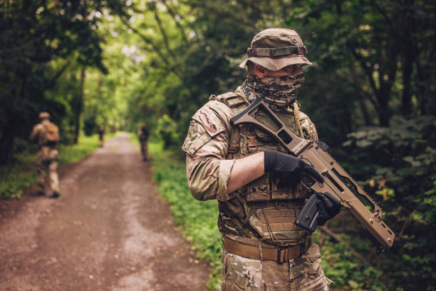 A tactical army personnel, dressed in camouflage uniform and carrying a gun, patrols along a forest trail, ensuring the region is secure and safe for navigation.