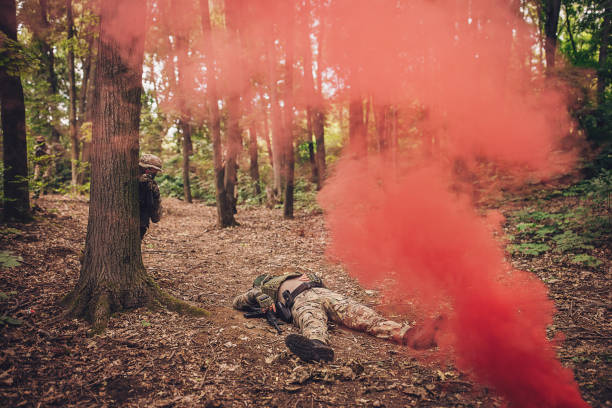 A soldier lies on the forest ground amidst red smoke, creating a dramatic and intense scene during a covert military operation, with others hiding nearby.