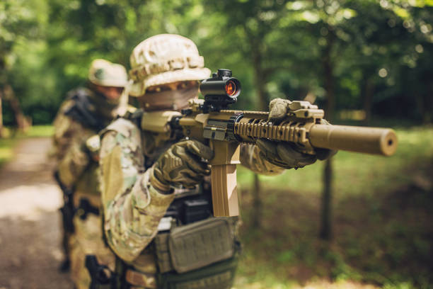 A soldier in full tactical gear and camouflage, aiming his rifle with precision while moving through a forest path, showing readiness and focus during a mission.