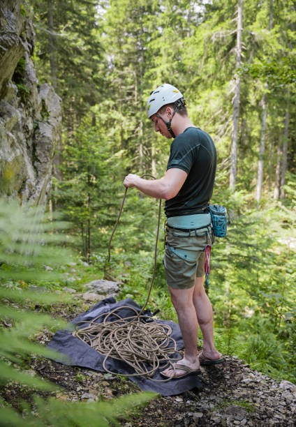 Man in climbing gear standing on rocky terrain with trees and a rope.