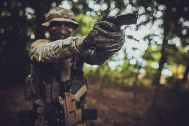 A heavily camouflaged soldier aims a pistol in a shaded forest, showcasing intense focus and readiness, with the natural wooded surroundings adding a sense of mystery and stealth.