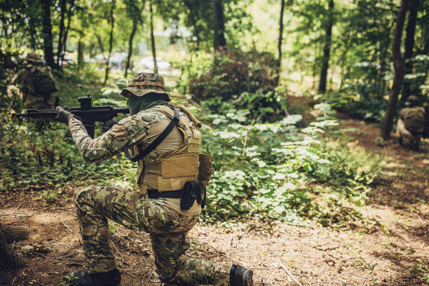 A military soldier in camouflage is seen kneeling in a forest setting during an exercise drill, holding a rifle and demonstrating high levels of readiness and strategic training.
