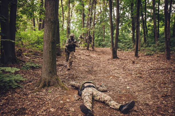 A soldier in full camouflage lies on the forest ground as another soldier approaches, depicting the harsh realities of military operations in challenging environments.